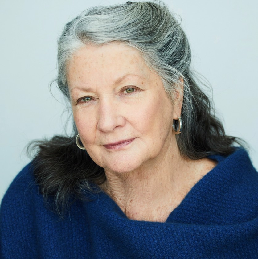 Close-up portrait of an older woman with silver hair and green eyes wearing a blue wrap in a brightly lit indoor environment.