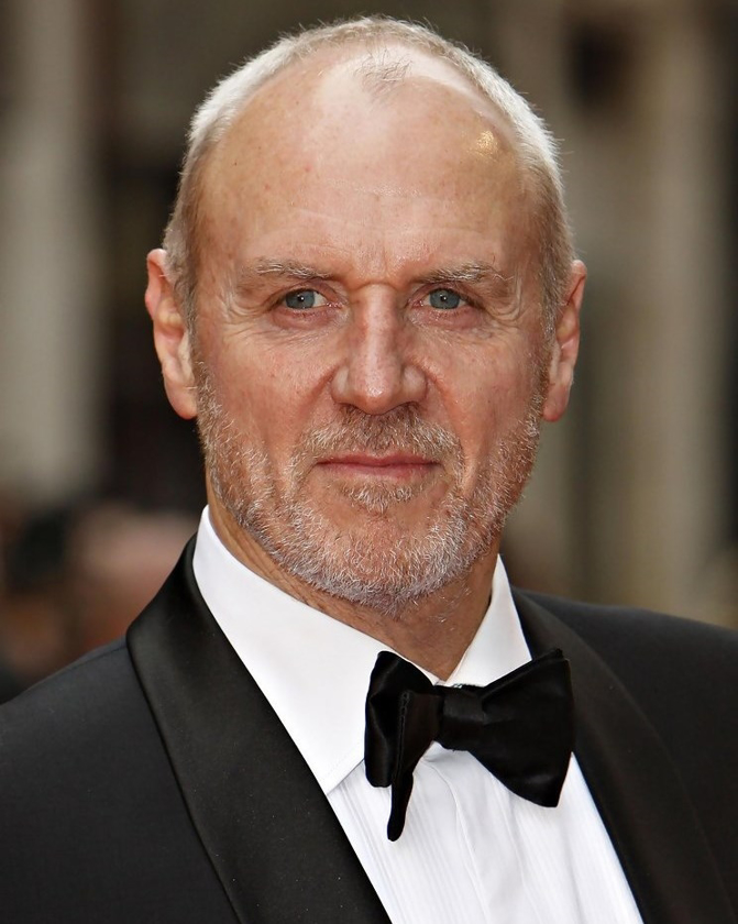 Close-up of an adult male with short white hair, wearing a black tuxedo and bow tie, against a neutral blurred background.