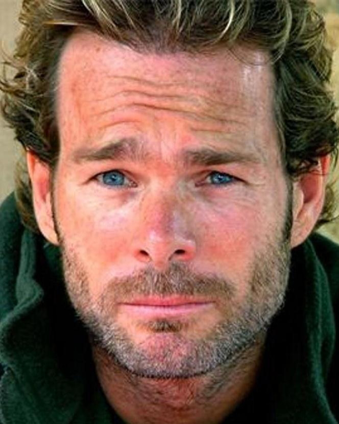 Close-up portrait of a fair-skinned adult male with light brown hair, stubble beard, and striking blue eyes looking directly at the camera.