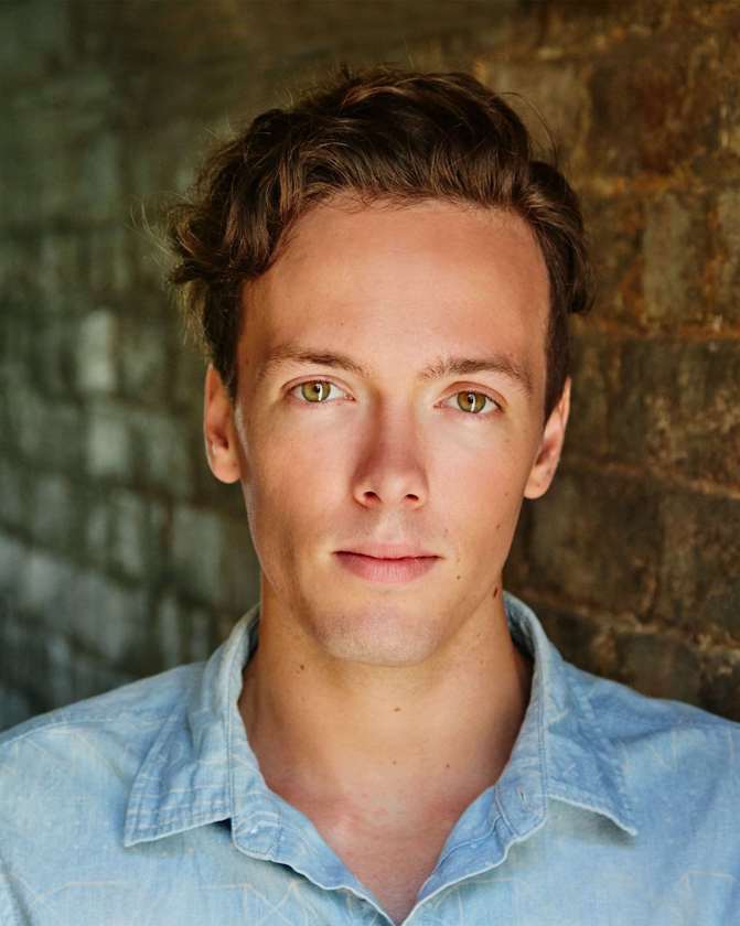 Close-up portrait of a smiling adult male with curly brown hair and green eyes wearing a light-blue button-down shirt, set against a brick wall.