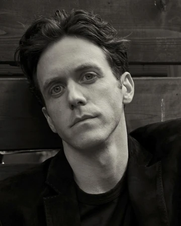 Portrait of a man in his 30s with dark wavy hair wearing a black jacket against a wooden wall in Sydney.