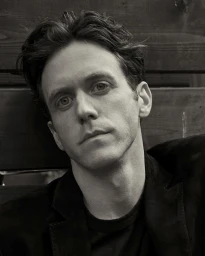 Portrait of a man in his 30s with dark wavy hair wearing a black jacket against a wooden wall in Sydney.
