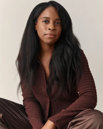 Portrait of a Black woman with long dark hair wearing a textured brown long-sleeve top against a neutral background.