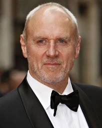 Portrait of an older man with short grey hair and facial stubble wearing a tuxedo with a black bow tie
