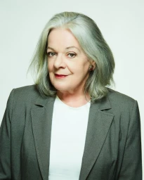 Portrait of an elderly woman with grey hair wearing a grey blazer and white top against a light background.
