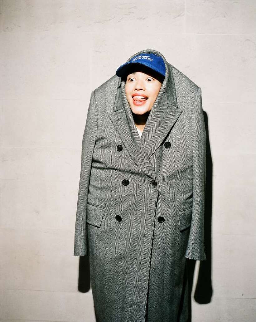A person is posing in a gray coat with a blue baseball cap, sticking their tongue out for the camera against a plain background indoors.