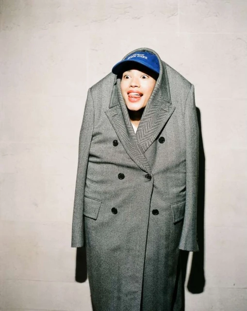 A person is posing in a gray coat with a blue baseball cap, sticking their tongue out for the camera against a plain background indoors.
