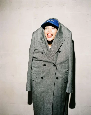 A person is posing in a gray coat with a blue baseball cap, sticking their tongue out for the camera against a plain background indoors.