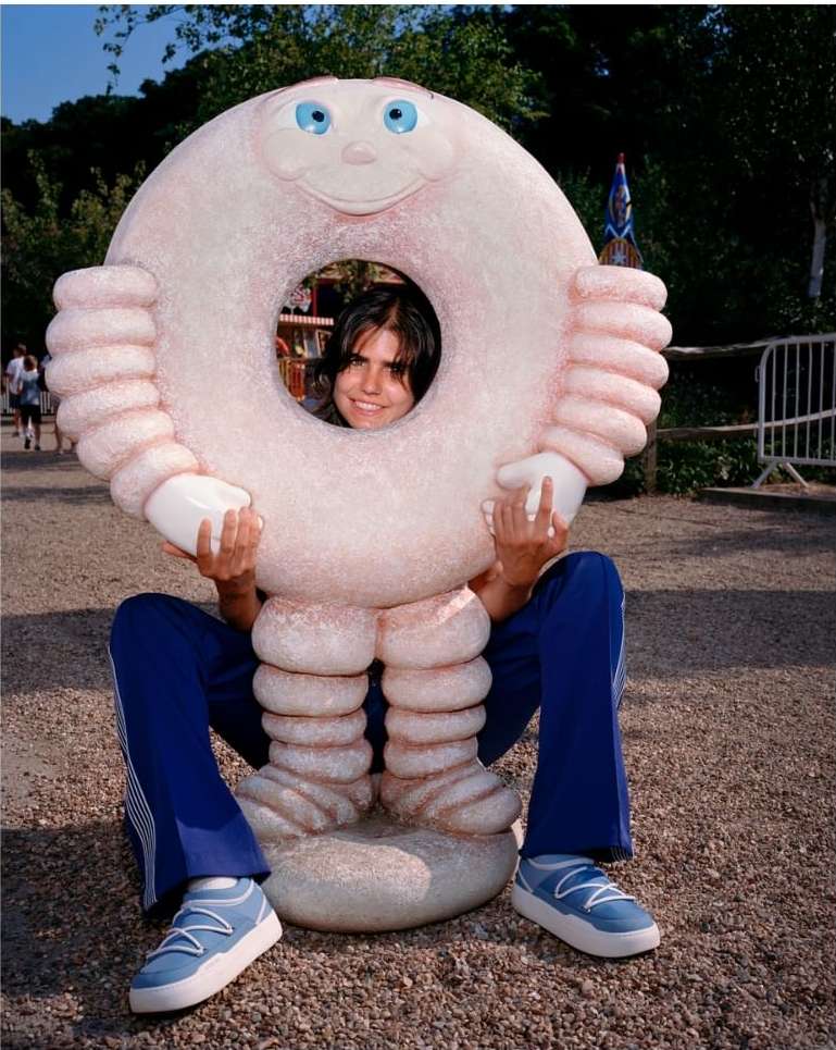 Person in a large novelty donut costume poses with a smile in an outdoor park setting on a sunny day, wearing casual blue clothing.