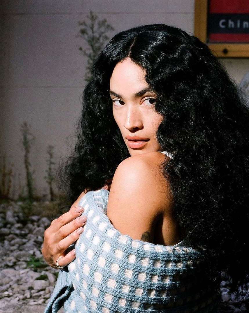 Portrait of a long-haired woman with dark hair partially wrapped in a checkered blanket looking over her shoulder outdoors.