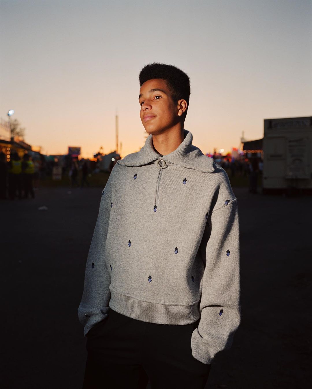 A young adult stands outside in a gray patterned quarter-zip sweater and black pants with an orange and blue sunset sky in the background.