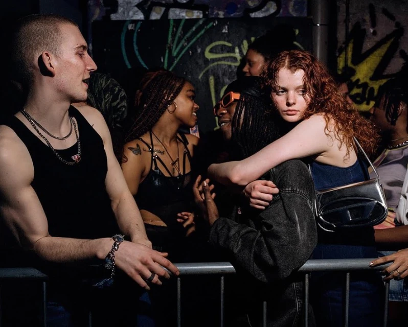 A group of young adults are standing in a dimly lit space in front of a metal railing, engaging in conversation with each other.