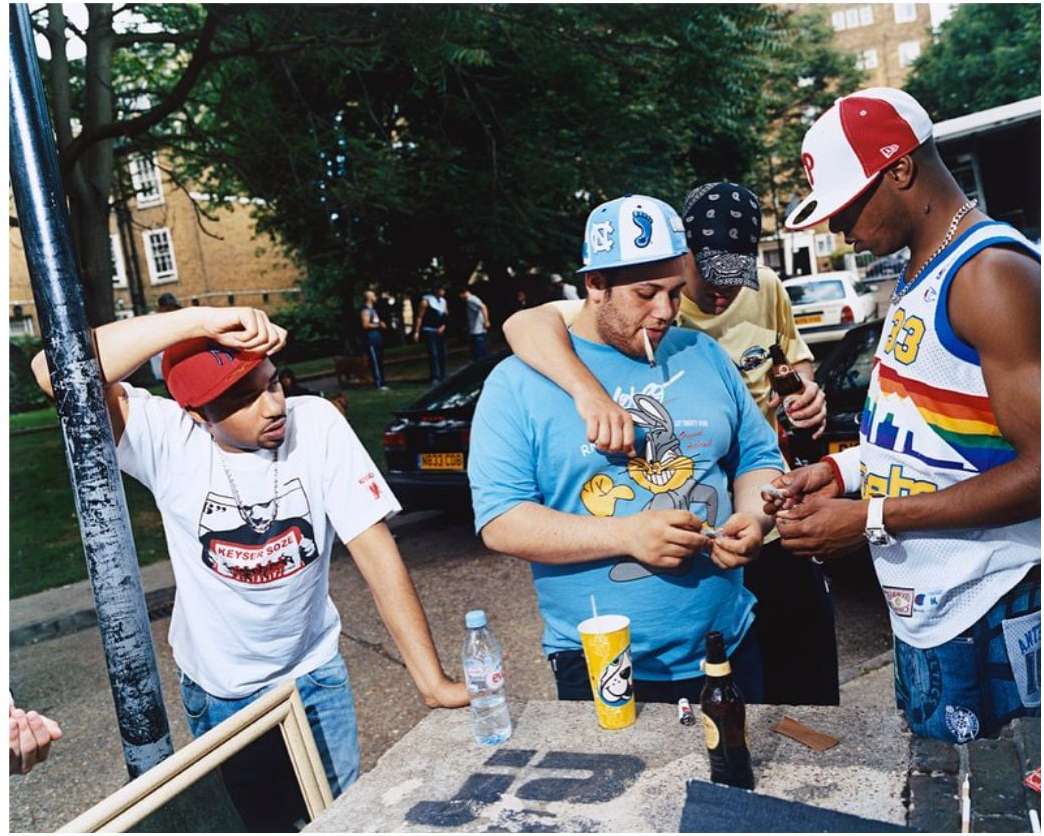 Group of casually dressed young men socializing outdoors, rolling cigarettes, with a residential street and cars in the background.