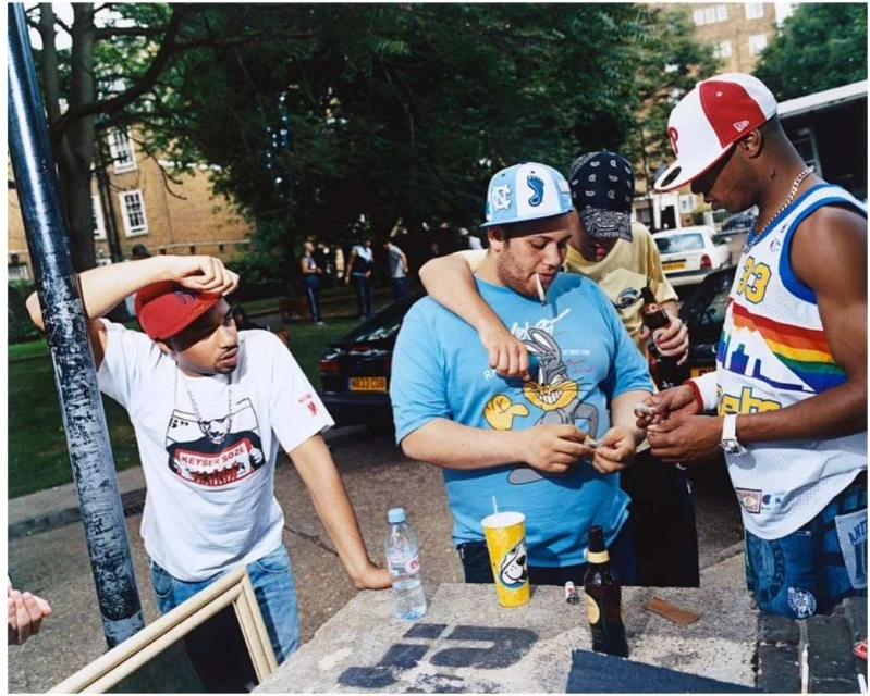 Group of casually dressed young men socializing outdoors, rolling cigarettes, with a residential street and cars in the background.