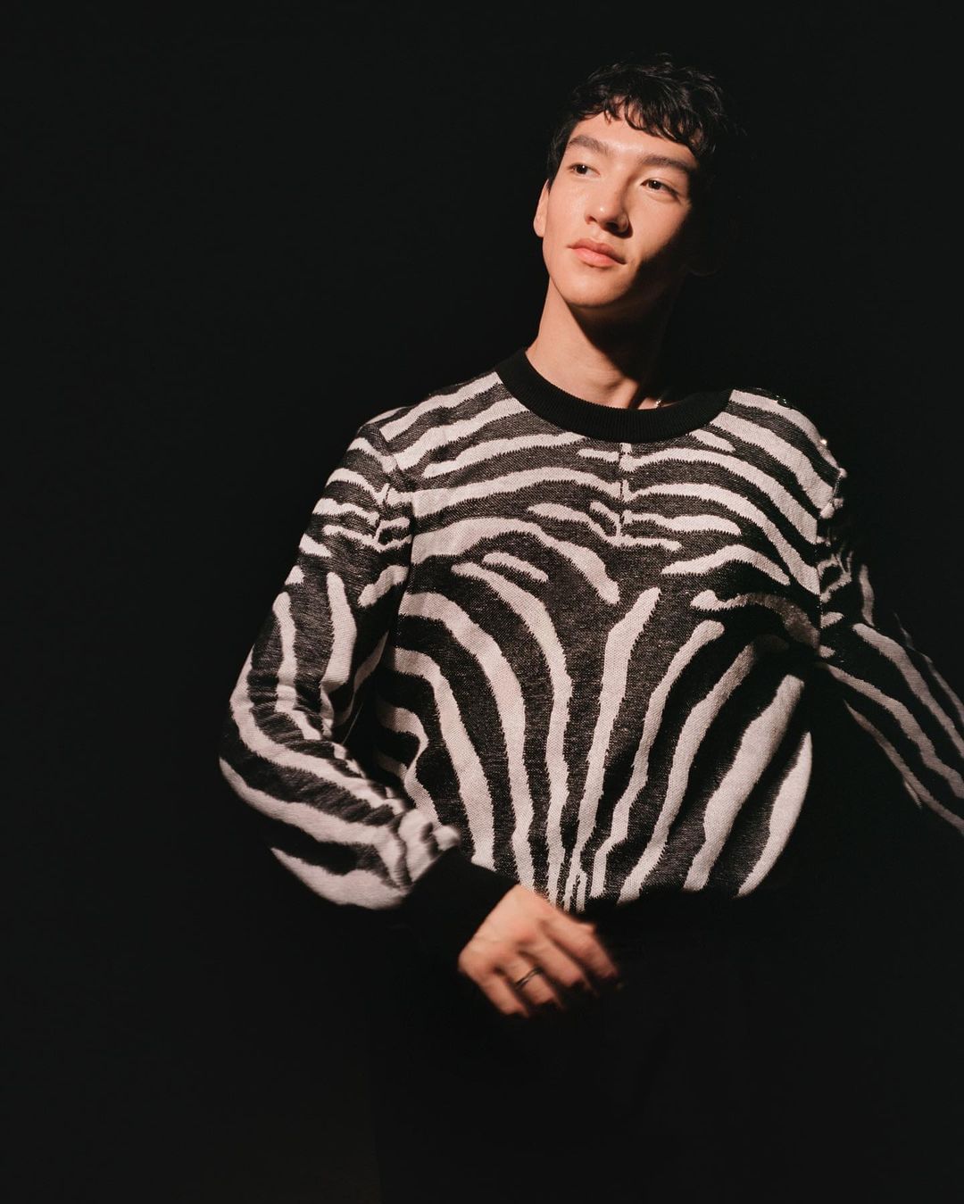 Adult male in zebra-striped sweater poses in a dimly lit photo studio with a black background and soft lighting. Fashion and portrait.