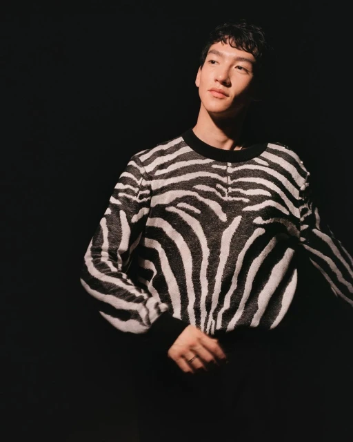 Adult male in zebra-striped sweater poses in a dimly lit photo studio with a black background and soft lighting. Fashion and portrait.