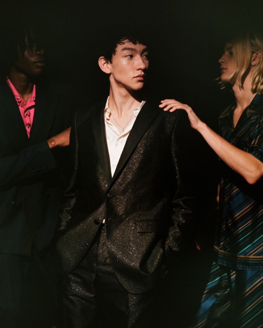 Three people pose in a dimly lit setting, with the focus on an Asian male wearing a sparkling black suit and white shirt.