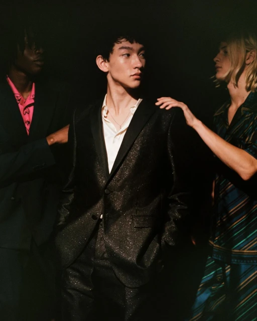 Three people pose in a dimly lit setting, with the focus on an Asian male wearing a sparkling black suit and white shirt.