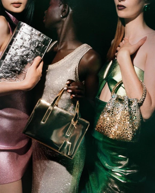 Three women pose indoors holding metallic handbags in pink, silver, and gold, wearing shiny dresses in front of a dark background.