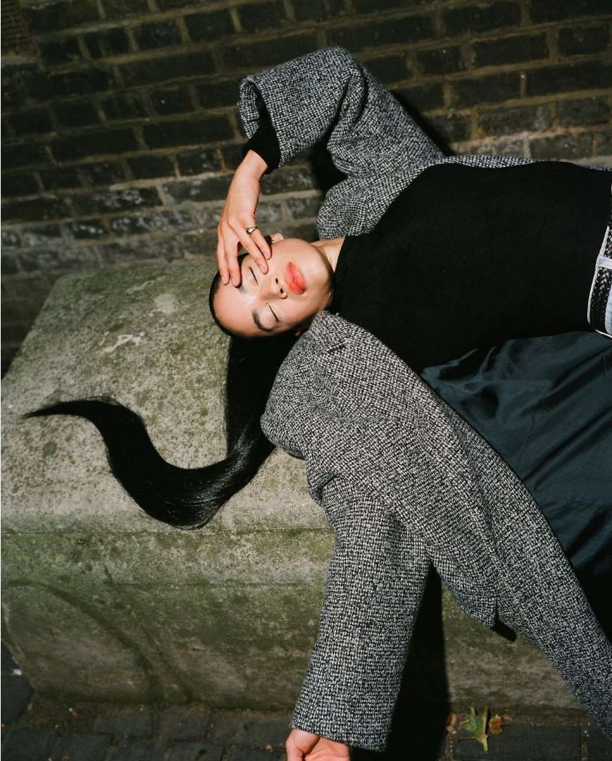 Model lying on stone block in grey tweed coat and black turtleneck with long dark hair