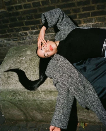 Model lying on stone block in grey tweed coat and black turtleneck with long dark hair