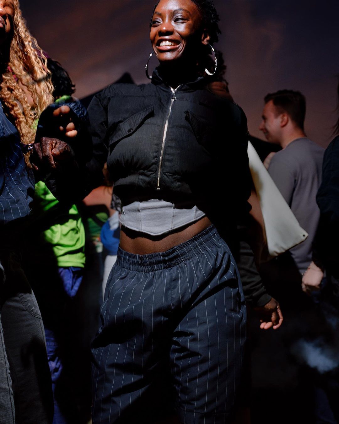 Smiling young woman dancing in a dark indoor space with other partygoers visible in the background, wearing a black jacket and striped pants.