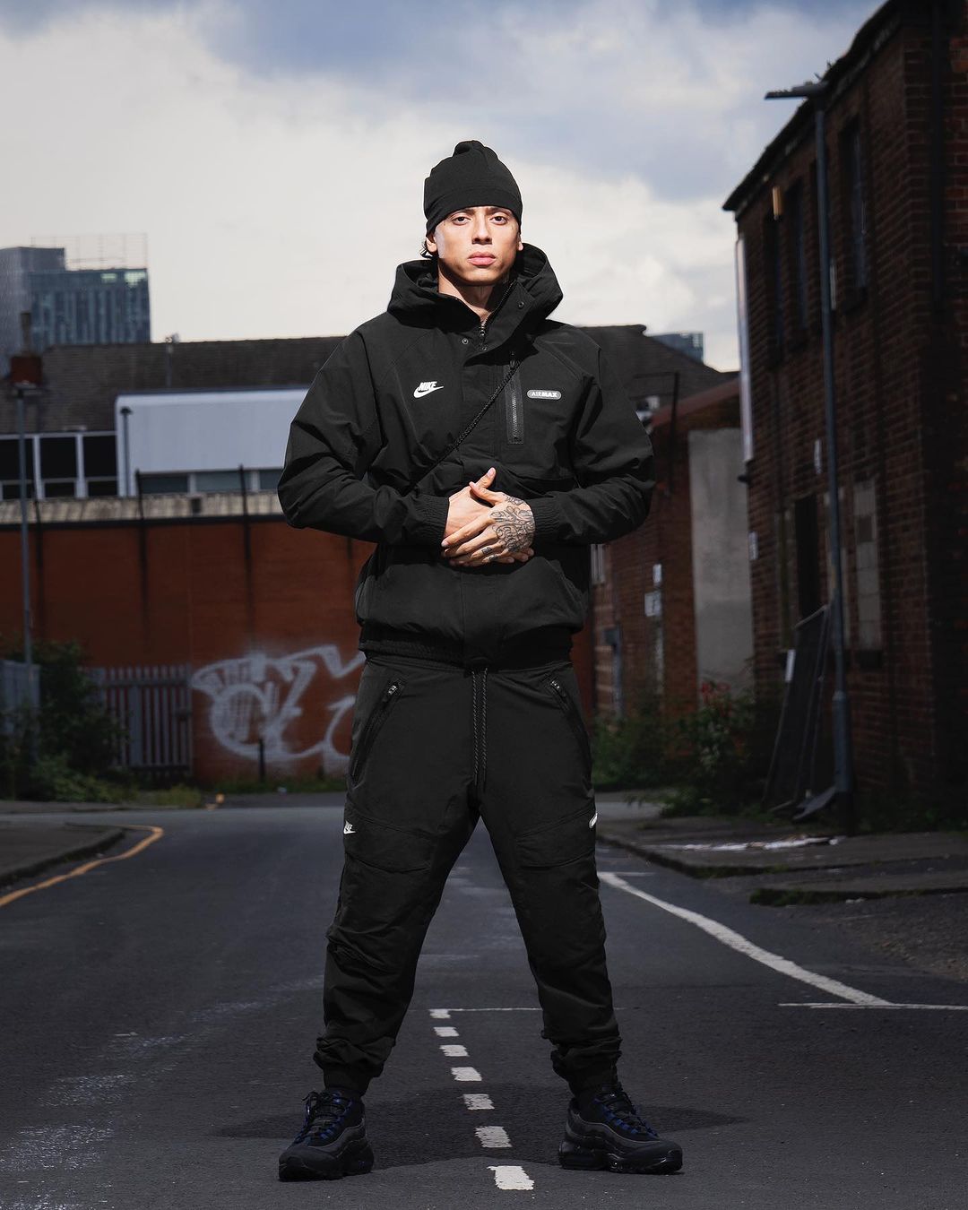 Man in black beanie, jacket, and pants poses on an empty street with buildings in the background. Full shot, urban environment.