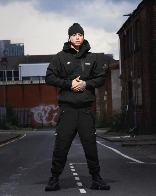 Man in black beanie, jacket, and pants poses on an empty street with buildings in the background. Full shot, urban environment.