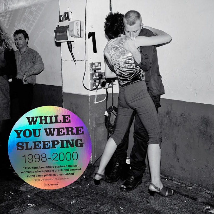 Black and white photo of a man and woman embracing and dancing in front of a wall with electrical outlets in an indoor setting.