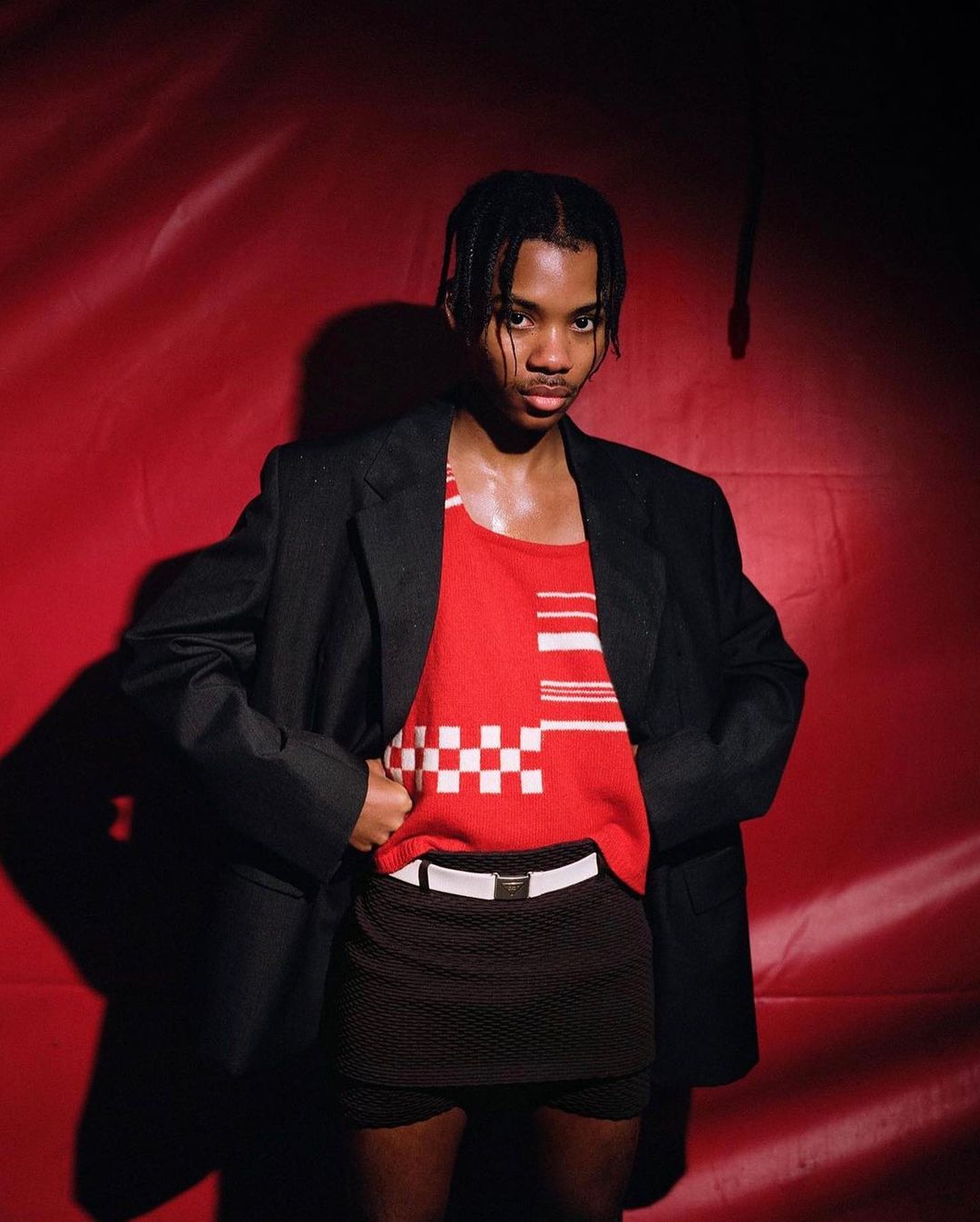 Adult male posing in red tank top, black jacket, and skirt against a red background in a studio setting under bright lighting.