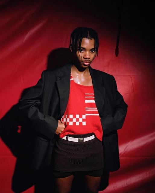 Adult male posing in red tank top, black jacket, and skirt against a red background in a studio setting under bright lighting.