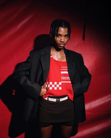 Adult male posing in red tank top, black jacket, and skirt against a red background in a studio setting under bright lighting.