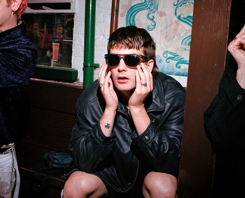 Young adult in sunglasses and leather jacket sits on a bench in an indoor environment, capturing a candid, urban, and fashion-forward moment.