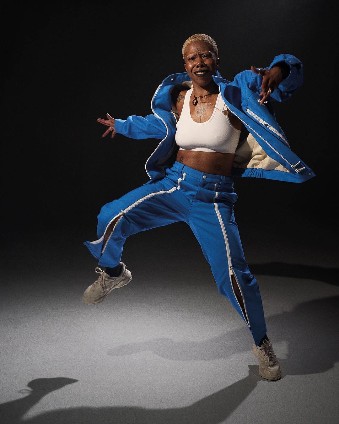 A mid-air shot of a short-haired Black woman in blue tracksuit with white top and tan shoes, set against a dark gray gradient background.