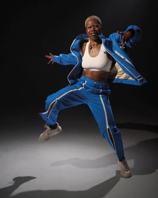 A mid-air shot of a short-haired Black woman in blue tracksuit with white top and tan shoes, set against a dark gray gradient background.