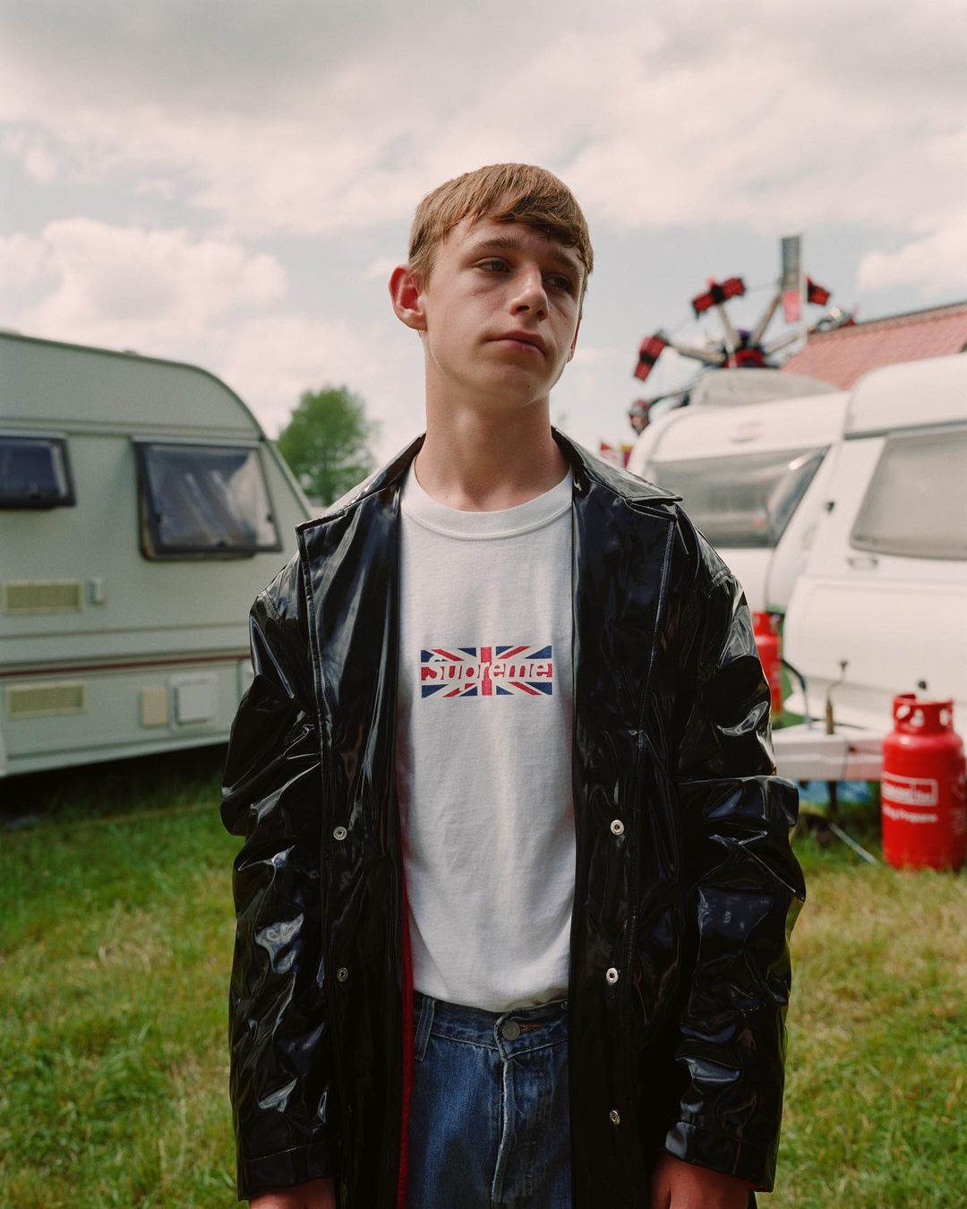 Teenager poses in black coat with logo t-shirt against a backdrop of travel trailers, grass and fairground rides on a cloudy day.