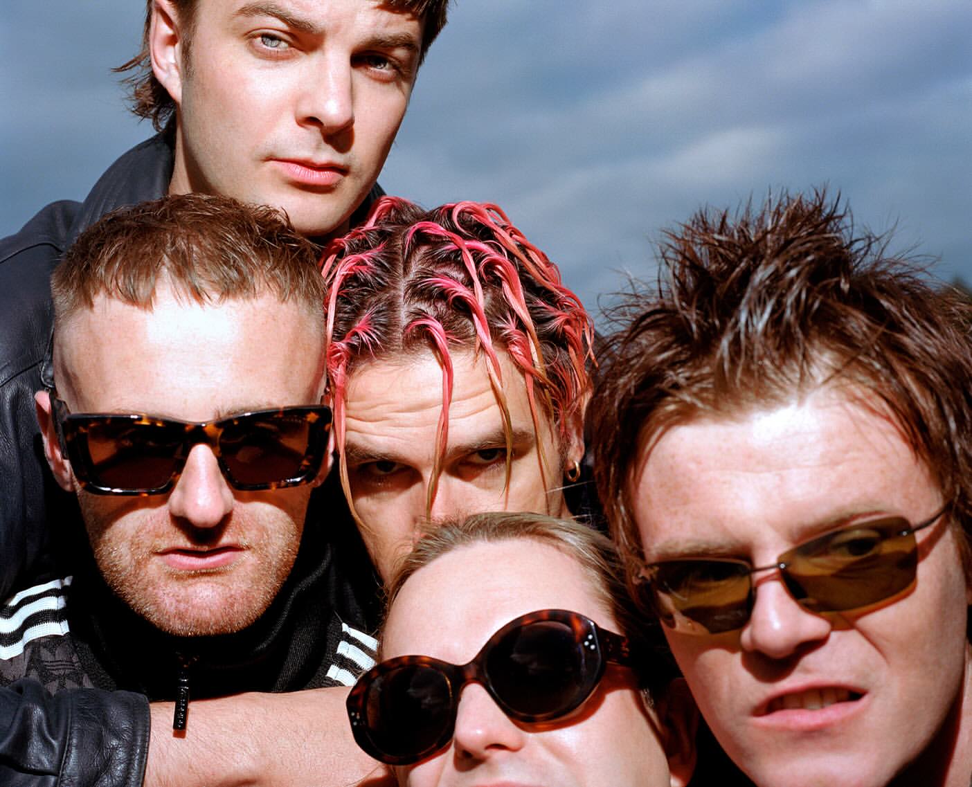 Close-up shot of five adult men wearing sunglasses outdoors, with some individuals sporting unique hairstyles and confident facial expressions.