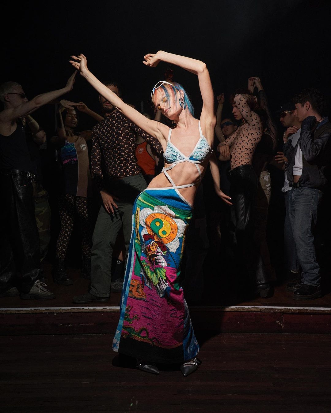 Woman dances in patterned swimsuit and wrap skirt at a crowded nightclub with colorful lights and other people dancing in background.