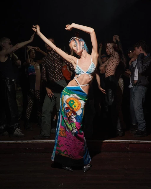 Woman dances in patterned swimsuit and wrap skirt at a crowded nightclub with colorful lights and other people dancing in background.