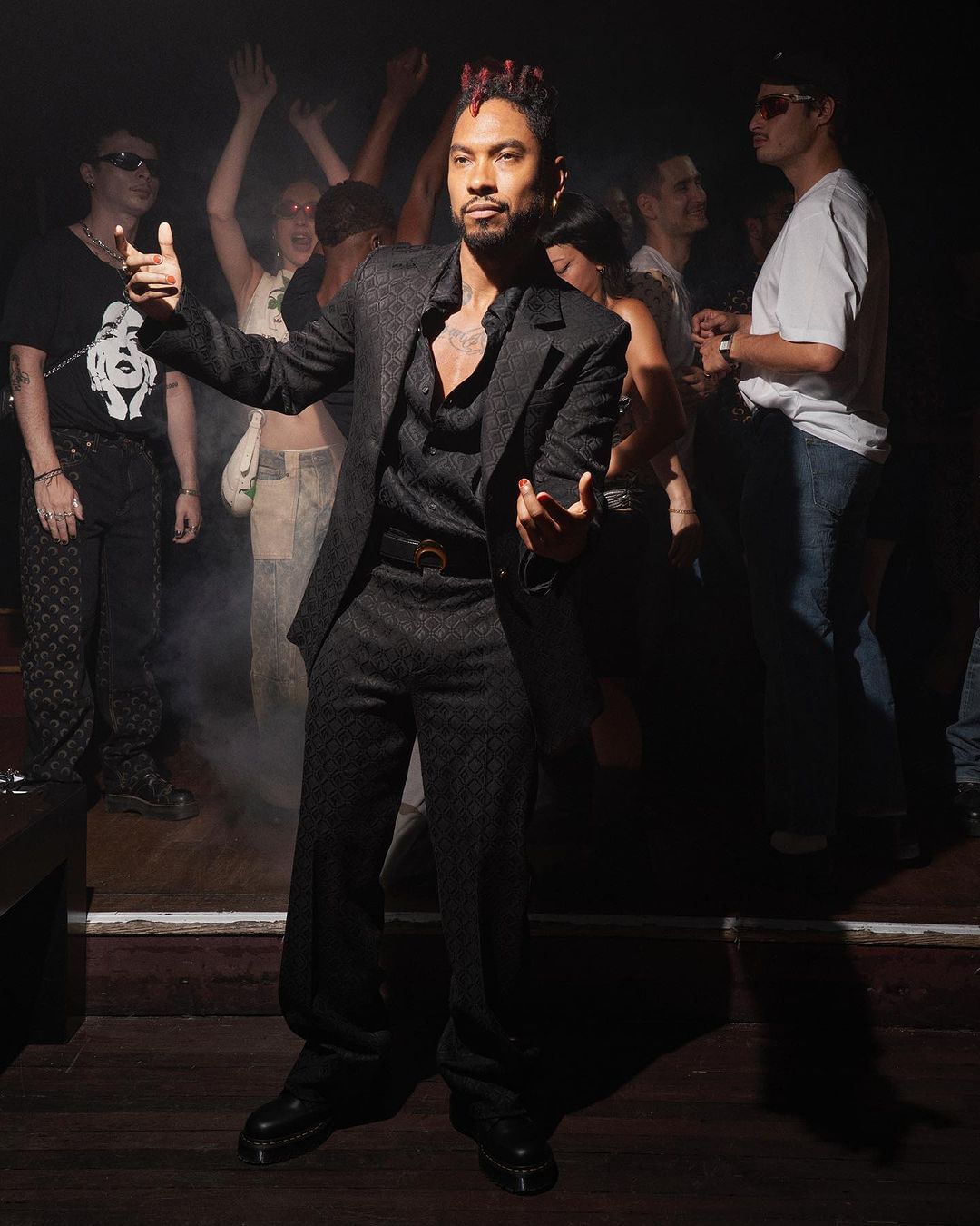Man in a dark suit stands confidently, gesturing in a dimly lit dance club with a crowd of people in the background, creating a vibrant atmosphere.