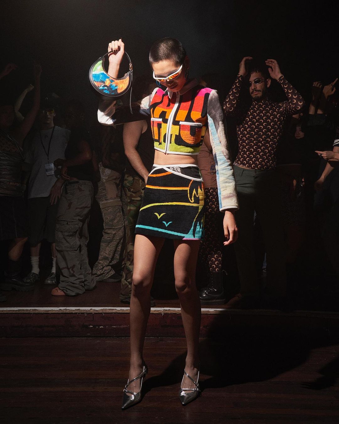 A fashion model on a dark stage wearing colorful, patterned, modern clothing with silver heels and holding a patterned bag. 