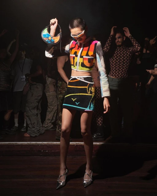 A fashion model on a dark stage wearing colorful, patterned, modern clothing with silver heels and holding a patterned bag. 