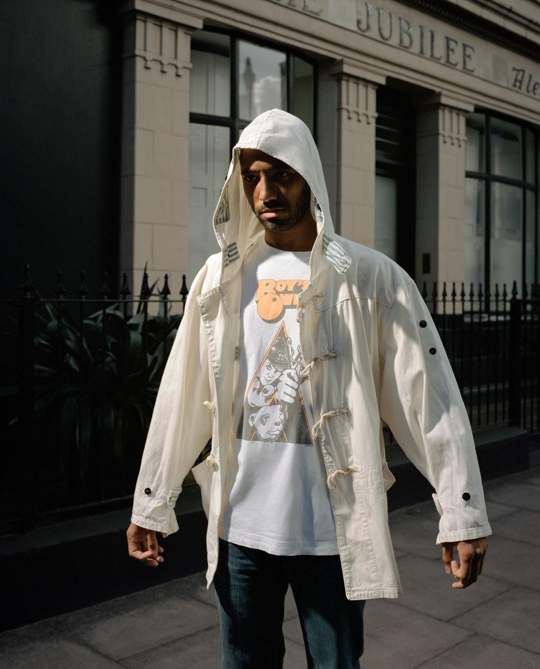 Man in hooded coat stands on urban sidewalk, wearing graphic tee and jeans, posing against building with classical architectural detail.