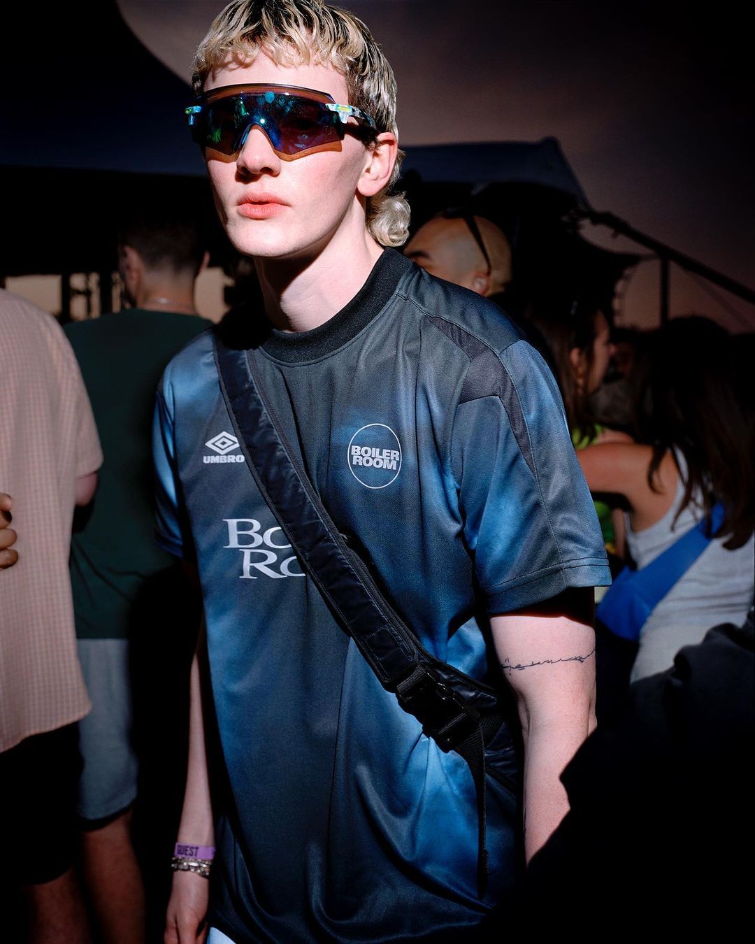 Close up of a young adult male wearing a blue soccer jersey, reflective sunglasses, and a black shoulder bag in a crowd of people.