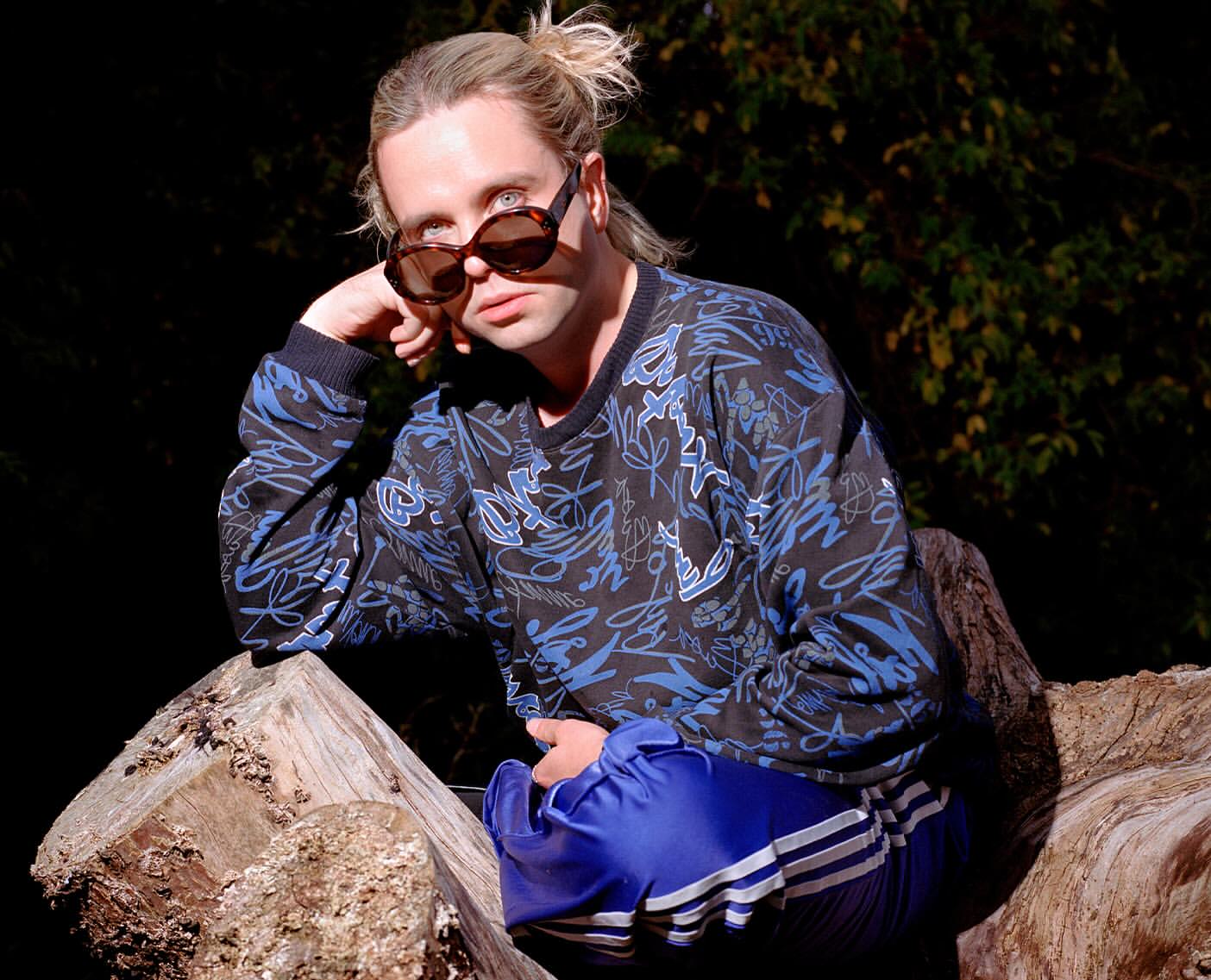 Portrait of a person wearing sunglasses and a patterned dark blue sweater while seated on a weathered tree trunk outdoors.
