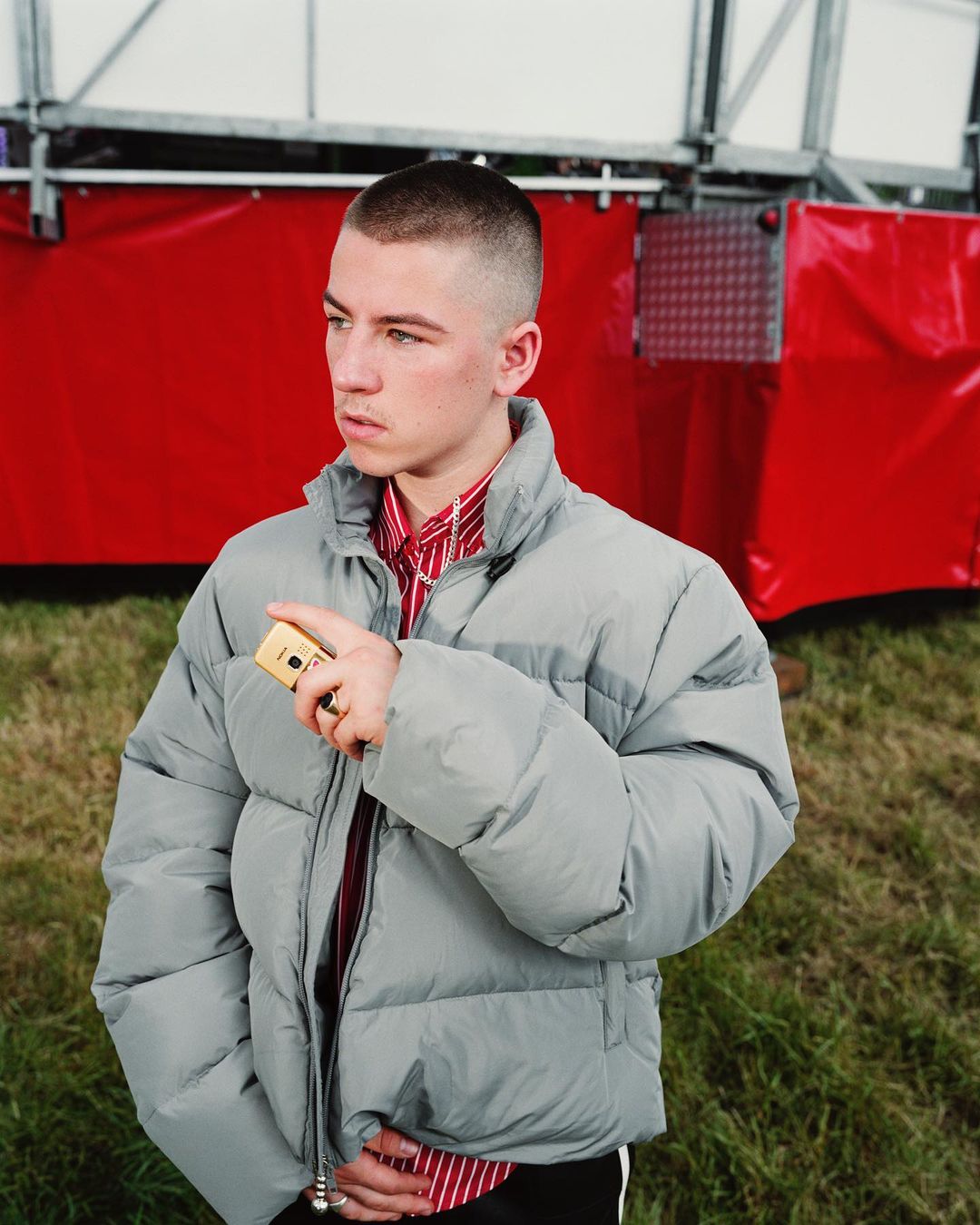 Young adult male in gray puffer jacket, red striped shirt, and silver chain poses outdoors with red and white structure behind him.