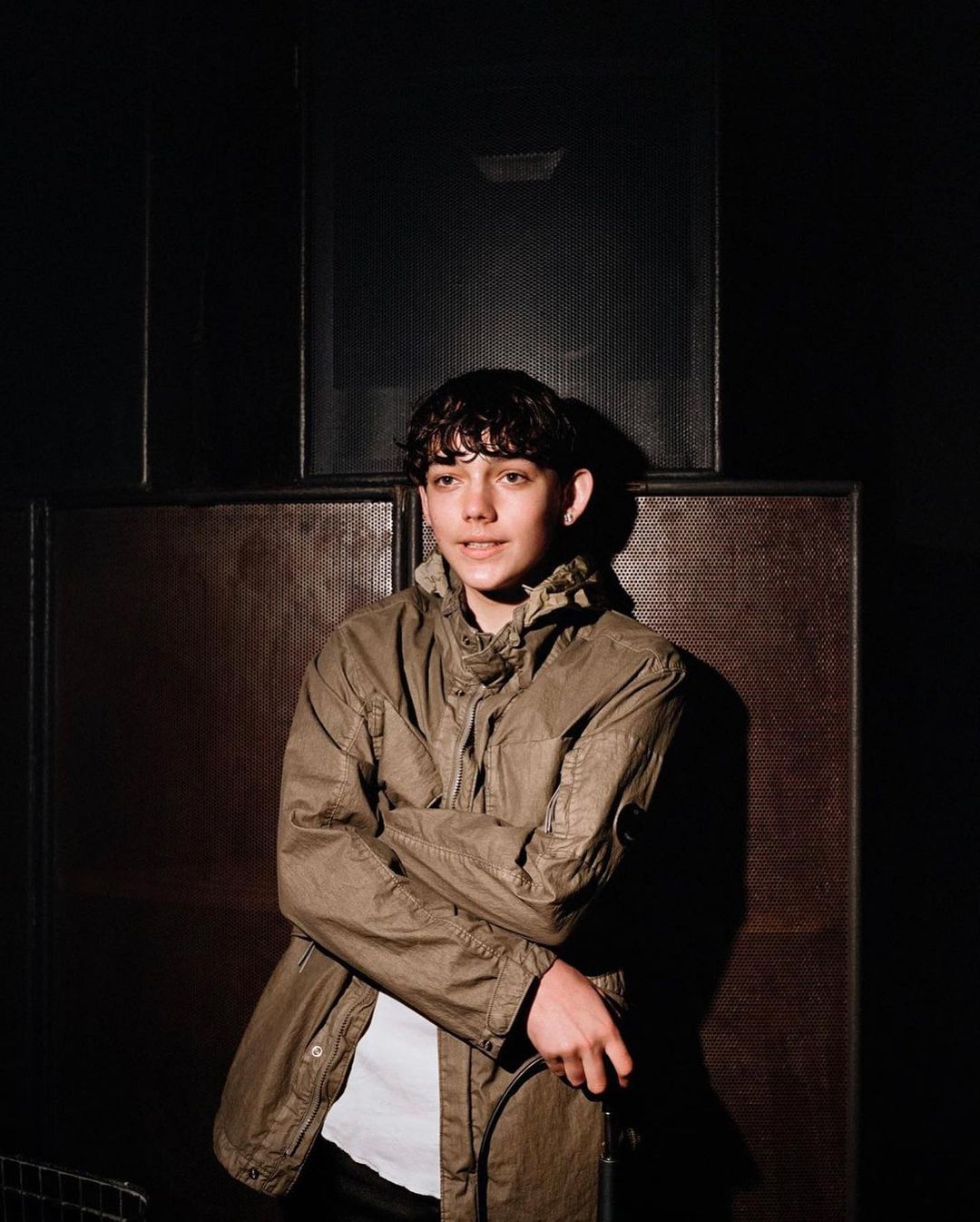 Portrait of a young man with curly hair in an olive-green coat, standing in a dimly lit room, looking at the camera with a slight smile.