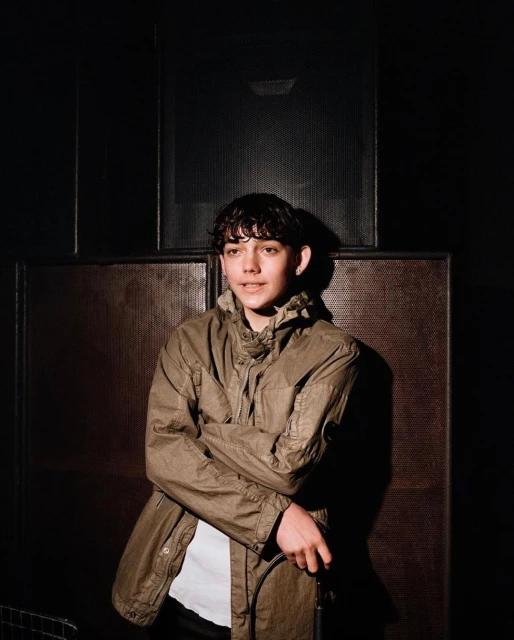 Portrait of a young man with curly hair in an olive-green coat, standing in a dimly lit room, looking at the camera with a slight smile.