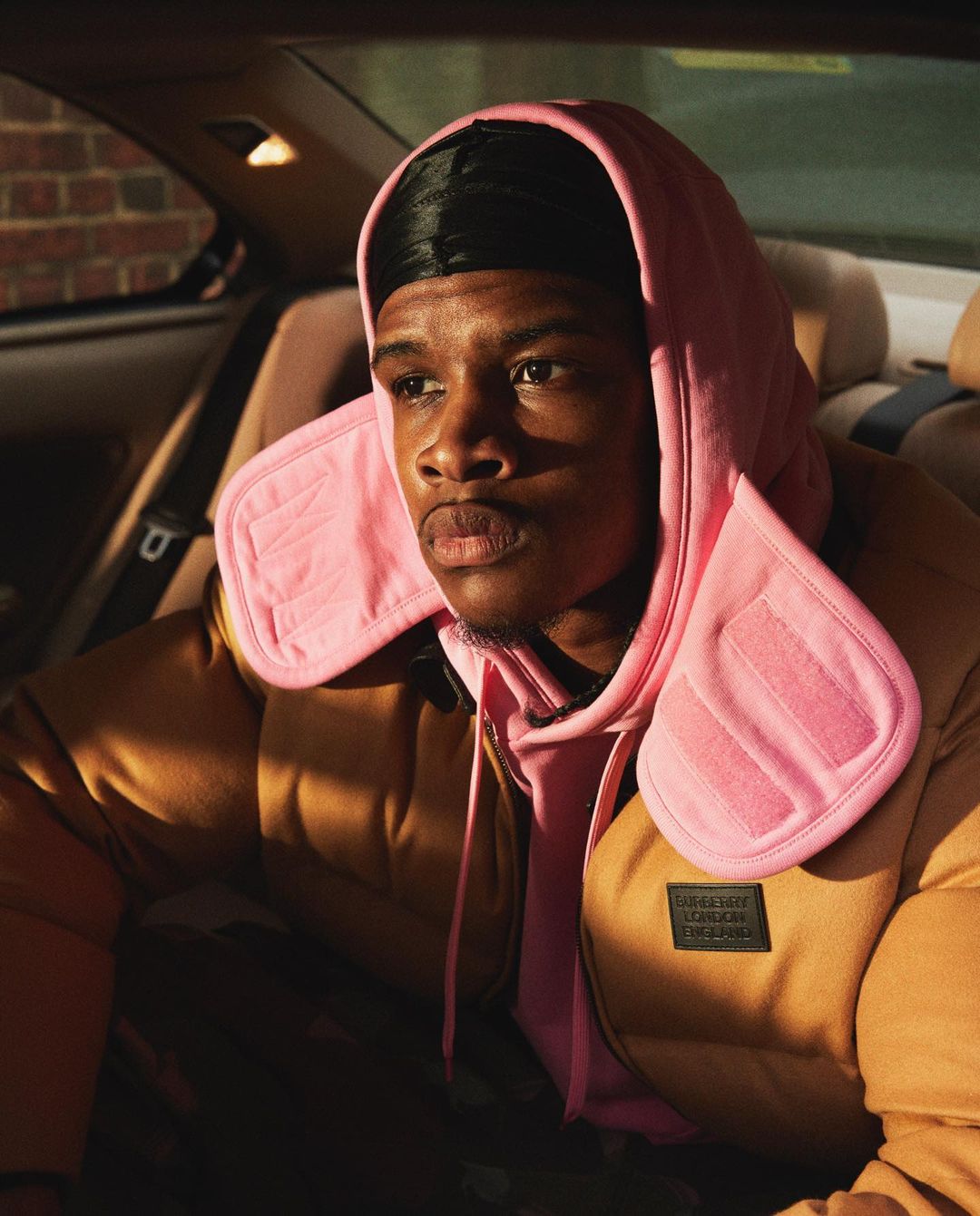 An adult male wearing a brown puffer jacket with a pink hoodie in a car, illuminated by natural light from the side window.