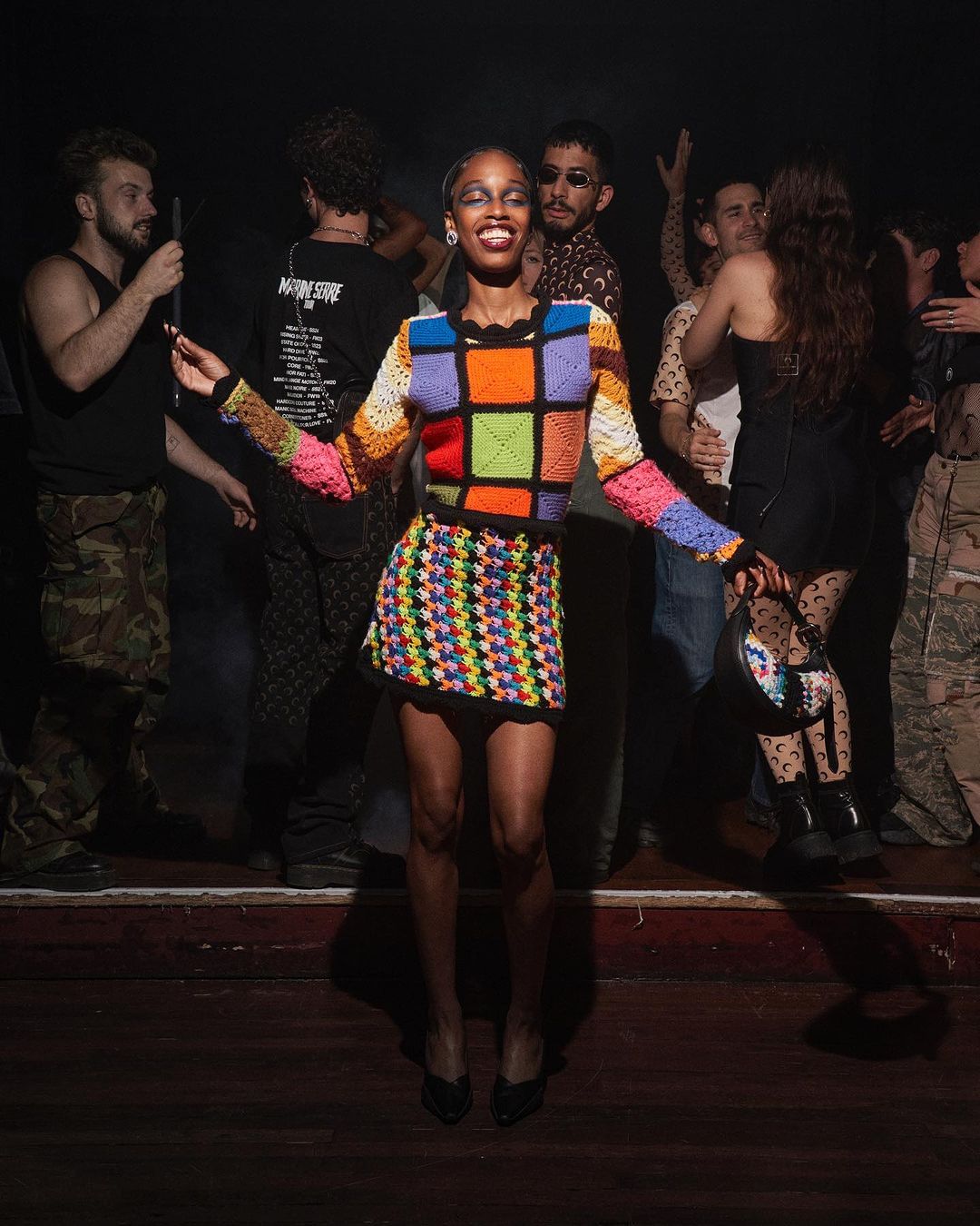 An adult woman in a colorful crochet sweater and skirt poses on a stage in front of a crowd of people in a dimly lit room.